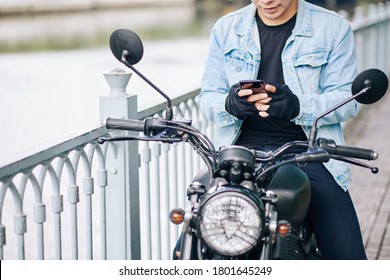 Cropped image of smiling biker in denim jacket and fingerless gloves sitting on motorcycle and texting friend or girlfriend
