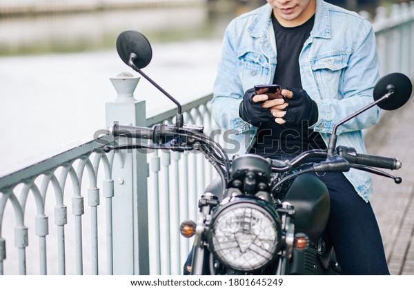Cropped image of smiling biker in denim jacket\
and fingerless gloves sitting on motorcycle and texting friend or\
girlfriend