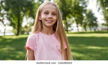 Cropped image of smiling caucasian girl looking at camera in blurred green park outdoors. Cute blonde female child of generation alpha wearing t-shirt. Concept of childhood lifestyle. Sunny summer day