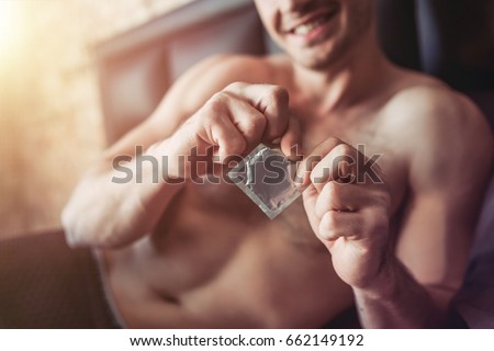 Cropped image of a smiling man holding condom in hand while lying on bed.