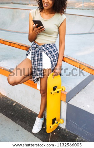 Cropped image of smiling young african girl using mobile phone while standing with skateboard outdoors