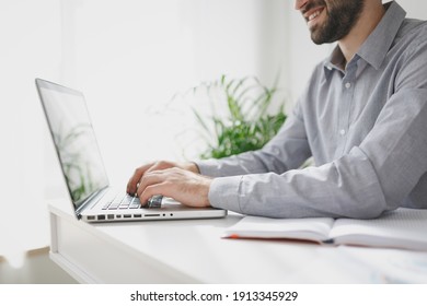 Cropped Image Of Smiling Young Bearded Business Man In Gray Shirt Sitting At Desk Working On Laptop Pc Computer In Light Office On White Wall Background. Achievement Business Career Concept