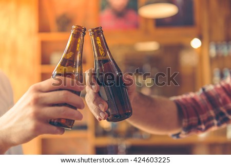 Cropped image of two men clanging bottles of beer together while sitting at bar counter in a modern urban cafe
