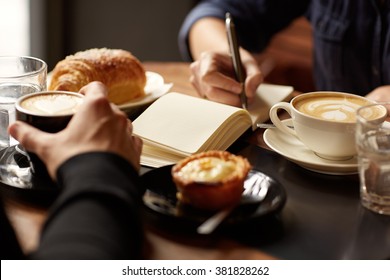 Cropped image of two people's hands at a table with coffees and pastry snacks, one person picking up their espresso while the other is writing in a notebook, possibly taking down an interview - Powered by Shutterstock