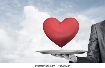 Cropped image of waitress's hand in white glove presenting big red heart on metal tray with cloudy skyscape on background.