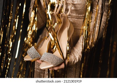 Cropped Image Of Woman In Gold Dress, Holding Golden Fancy Fashion Jewelry Shoes High Heels, Open-toed With Soft Inner Sole. Preparing For Prom Ballroom Party Celebration. Gold Lametta On Background.