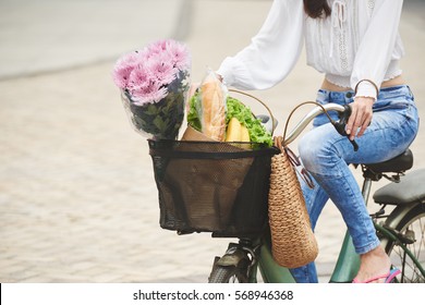 Cropped Image Of Woman Riding Bicycle With Basket Of Grocery And Flowers