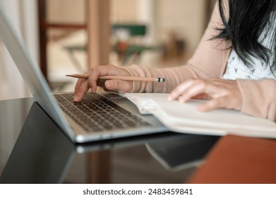 A cropped image of a woman working on her laptop computer, taking notes in a book and typing on the laptop keyboard at a table indoors or in a cafe. people and technology concepts