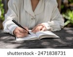 A cropped image of a woman writing something in a book or keeping her diary at a wooden table in a garden on a bright day. people and lifestyle concepts