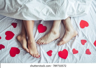 Cropped image of young couple is lying on bed. Close up of male and female feet. Loving couple is lying on bed under blanket covered by small red paper hearts. Saint Valentines Day.