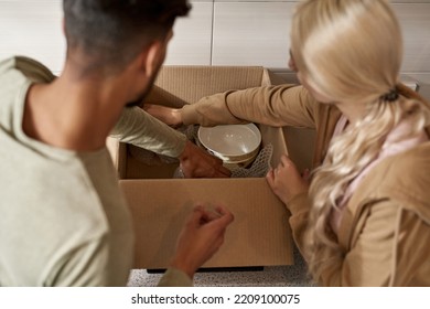 Cropped Image Of Young Couple Packing Cooking Utensils In Air Bubble Film In Cardboard Box At Kitchen. Blonde Girl With Tattoo And Bearded Man Moving To New Apartment. Concept Of Home Relocation