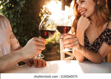 Cropped image of young loving couple sitting in cafe by dating outdors in park holding glasses of wine drinking.