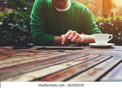 Cropped image young man sitting at the table with cup of coffee, digital tablet and smart phone, man's hands typing message on touch screen tablet while sitting on balcony terrace, flare sun