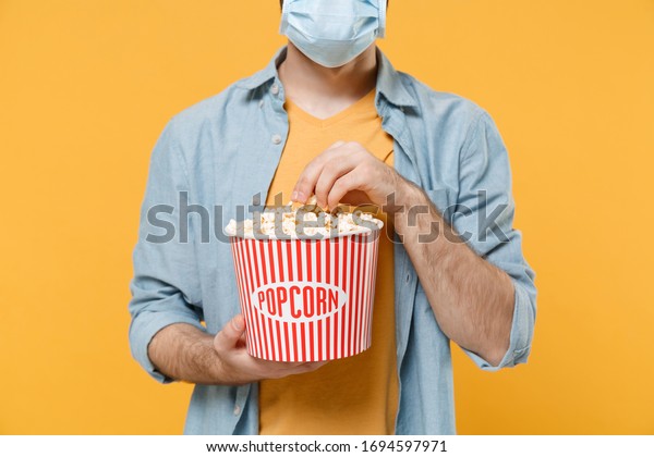 Cropped image of young man in sterile face mask isolated on yellow wall background. Epidemic pandemic coronavirus 2019-ncov sars covid-19 flu virus concept. Watching movie film hold bucket of popcorn
