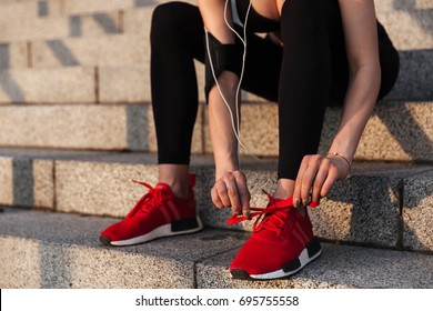 Cropped image of young sports lady sitting with earphones outdoors and tie laces.