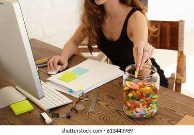 Cropped image of young stressed woman eating sweets at workplace in office. The girl takes candy from big glass jar with lollipops standing on a desktop. Stress and junk food concept