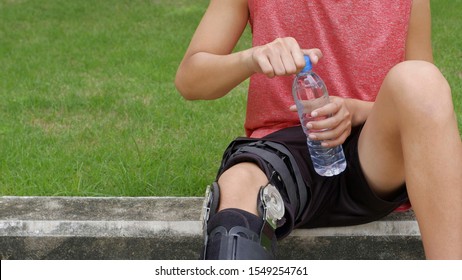 Cropped Image Of Young Teenager Boy Wears Supportive Knee Brace, Relaxing And Open Fresh Water Bottle To Drink During Jogging In Public Park At Morning Time, Exercise And Health Care Concept 