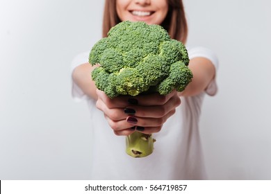 Cropped image of young woman dressed in white t-shirt showing broccoli to camera.