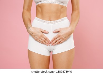 Cropped image of young woman wearing white brassiere underwear with sports fit body posing hands touching gesturing pain in stomach intimate zone isolated on pastel pink background, studio portrait