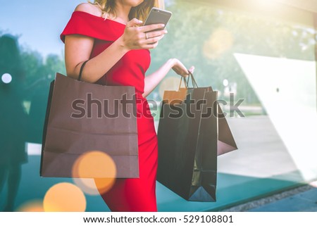 Cropped image.Young smiling woman with blond hair,dressed in red fitting dress,standing outside with shopping bags and using smartphone.Girl uses digital gadget. Summer day.On background glass wall.