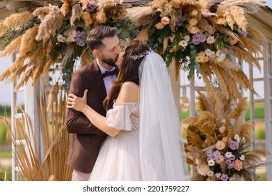 Cropped of loving brides couple, elegant bride with veil, wearing in wedding dress kissing with stylish groom in brown suit while standing together near lake and decorated with dried flowers gazebo - Shutterstock ID 2201759227