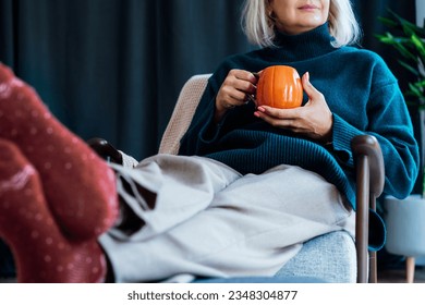 Cropped mature middle aged woman in sweater and warm socks relaxing in armchair with pumpkin shaped cup of hot coffee or tea drink. Cozy calm autumn holidays at home. Fall hygge mood concept.