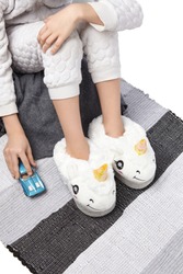 Cropped Medium Shot Of A Child In White Romper With Texture Pattern And In White Plush House Slippers Made As Lamb Unicorn. The Child With A Blue Car Is Sitting On The Striped Carpet And A Gray Plaid.
