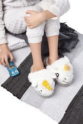 Cropped Medium Shot Of A Child In White Textured Pyjamas And Plush House Slippers Made As White Smiling Lamb Unicorn. The Child With A Blue Car Is Sitting On The Striped Carpet And A Gray Plaid