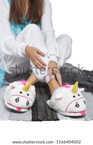 Cropped medium shot of a lady in white velour pyjamas and plush house slippers made in the form of white teasing unicorn. The girl is sitting on the striped carpet and a gray plaid.