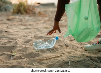 Cropped photo of African woman collecting spilled trash garbage from sand on beach in green plastic bag. Womans hand cleaning up used plastic bottles. Ecology, environmental conservation, pollution.  - Powered by Shutterstock