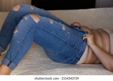 Cropped photo of barefoot fat plump plus-size overweight woman lying on beige plaid, wearing nude bra and trying to pull on blue ripped torn jeans indoors. Body positive, cellulite, unhealthy diet.