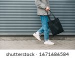 Cropped photo of the bottom of a young man in stylish clothes walking on the background of a gray wall with an black eco bag in his hand. Eco friendly. Reusable bag in young man