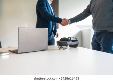 Cropped photo of a business hand shake next to laptop and augmented mixed reality goggles - Powered by Shutterstock