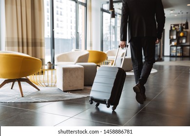 Cropped photo of caucasian businessman wearing formal black suit walking with suitcase in hotel lobby