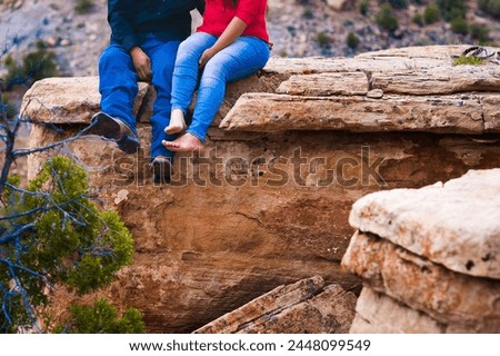Cropped photo of a couple as they sit together with their feet and legs dangling off a small cliff. They are amongst the red rocks of the Bears Ears National Monument.