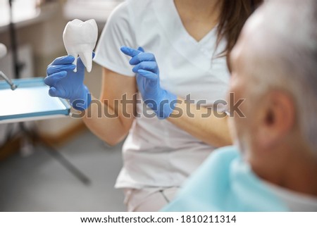 Cropped photo of dentist holding a big white model of a tooth while showing it to a senior patient