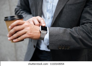 Cropped photo of european businessman in gray suit and white shirt standing against granite wall and touching smartwatch while drinking takeaway coffee