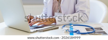 Cropped photo of female doctor in white uniform working on laptop computer in hospital, sitting at desk with stetoscope and ultrasound results. Medical panoramic banner for website header