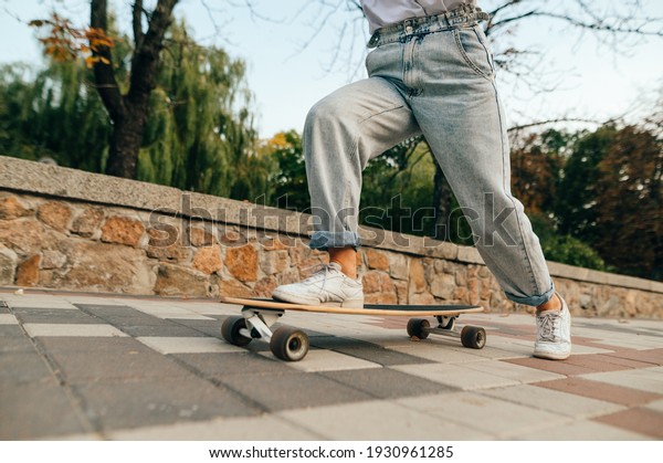 Cropped photo of\
female skateboarder legs in baggy jeans, riding her long board on a\
city street near the\
park.