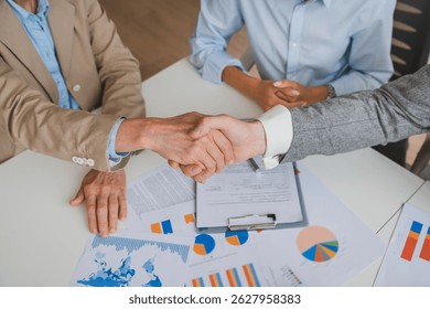 Cropped photo of handshake in agreement against young business people in board room meeting discussing analyzing graphs financial data and planning a marketing project together.