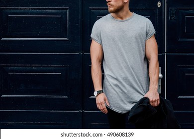 A cropped photo of a handsome muscular man wearing blank gray t-shirt while standing beside the dark blue entrance door on a street. Empty place for the logo or design. Mock up.