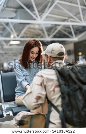 Cropped photo of happy female model looking down while sitting and holding her husband's arms indoors. American soldier is situating in front of his spouse. Homecoming concept
