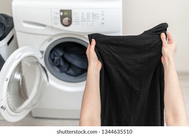 Cropped Photo Of Laundry Process. Woman Holding Black Clothes In Her Hands Against White Washing Machine Inside Light Flat Interior