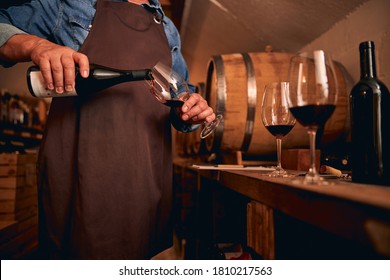 Cropped photo of a male sommelier in a brown apron standing in the cellar and pouring wine into a glass