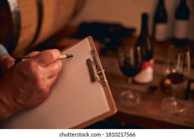 Cropped photo of a man holding a clipboard and a touching a sheet of paper with a pen. Glasses and bottles of red wine on the background - Powered by Shutterstock