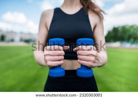 Cropped photo of a slender athletic young girl who goes in for sports in the stadium with dumbbells. Athletic woman with dumbbells outdoors.