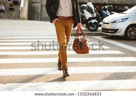 Cropped photo of stylish successful man in business wear walking through pedestrian crossing in downtown with leather male bag in hand
