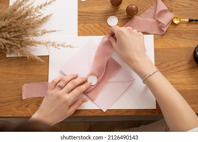 Cropped Photo Woman Hands Making Greeting Envelope Craft, Decorating With Pink Ribbon And Stamping. Cereals On Desk. 