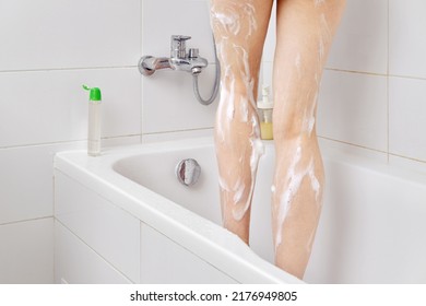 Cropped Photo Of Woman Using Showering Gel While Taking Shower In Bathroom, Standing In Bathtub. Close Up Of Slim Female Legs Covered With Soap Suds Or Foam. Hygiene And Body Wash Concept