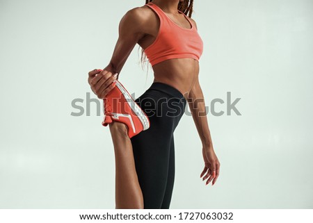Cropped photo of young african woman with perfect body stretching her leg while standing against grey background